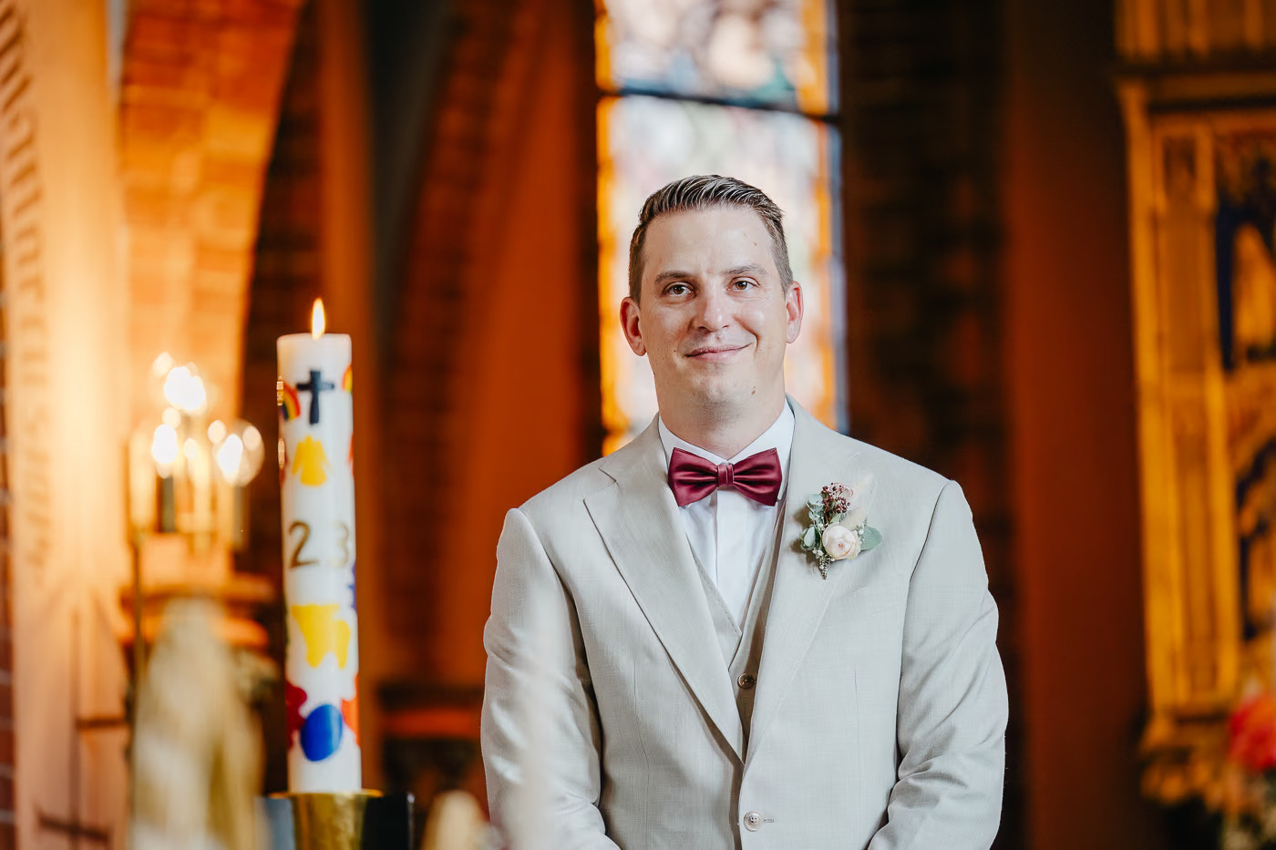 Kirchliche Trauung Helmstedt – Bräutigam im hellen Anzug mit Fliege wartet lächelnd am Altar, Hochzeitskerze mit Kreuz im Vordergrund, emotionale Hochzeitsfotografie Helmstedt Kathrin Skorpel