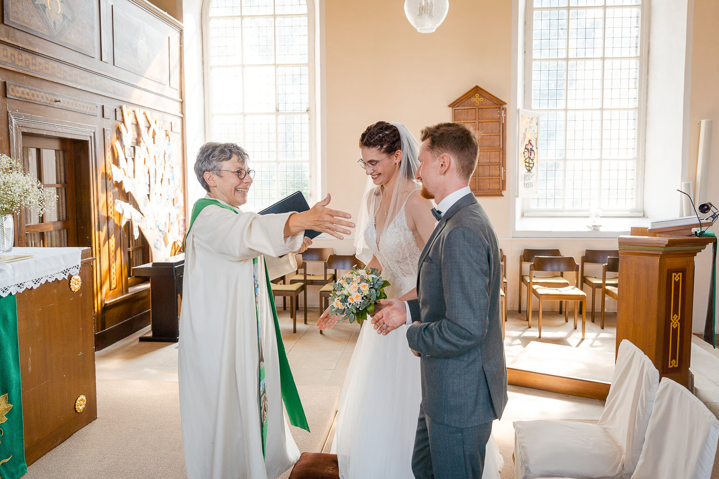 Kirchliche Trauung Helmstedt – Pfarrerin begrüßt das Brautpaar lachend mit offenen Armen in der Kirche, warme Atmosphäre, kirchliche Hochzeitsfotografie Helmstedt Kathrin Skorpel