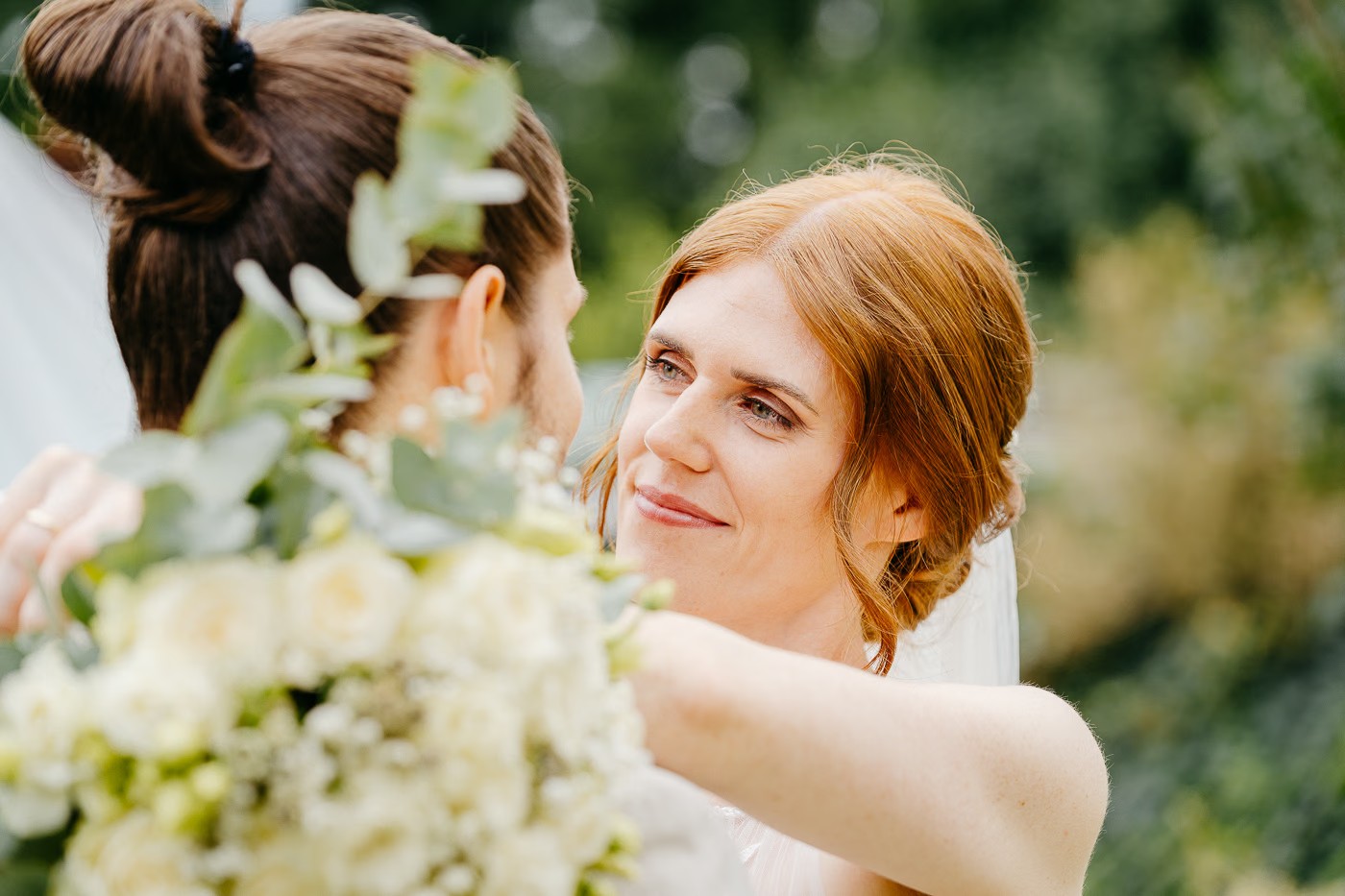 Paarshooting Gifhorn – Braut hält lächelnd einen Strauß weißer Rosen zwischen sich und dem Bräutigam mit Dutt, grüner Blätterhintergrund, verspielte Nähe, Hochzeitsfotografie Gifhorn Kathrin Skorpel