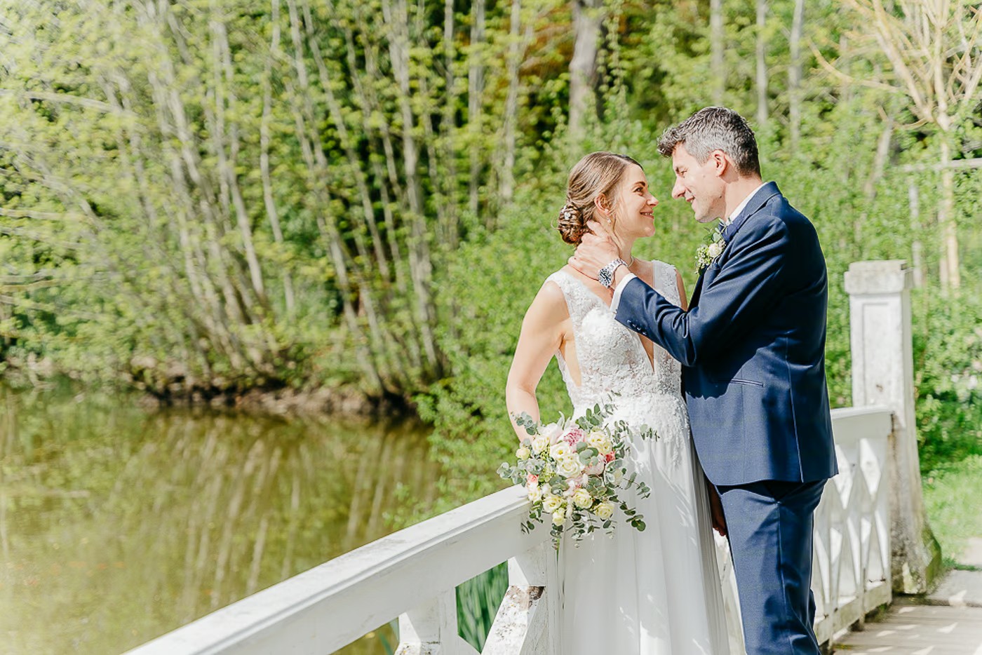 Paarshooting Schlosspark Cremlingen – Brautpaar steht auf der weißen Holzbrücke über den Teich, Bräutigam hält zärtlich den Nacken der Braut, Wasserspiegelung im Hintergrund, Hochzeitsfotografie Schlosspark Cremlingen Kathrin Skorpel