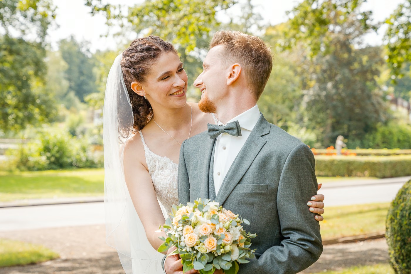 Paarshooting Bad Helmstedt – Brautpaar blickt sich lächelnd in die Augen, Braut mit Flechtfrisur und Schleier hält Rosenstrauß in Apricot, Parkweg mit Hecken im Hintergrund, herzliche Vertrautheit, Hochzeitsfotografie Bad Helmstedt Kathrin Skorpel