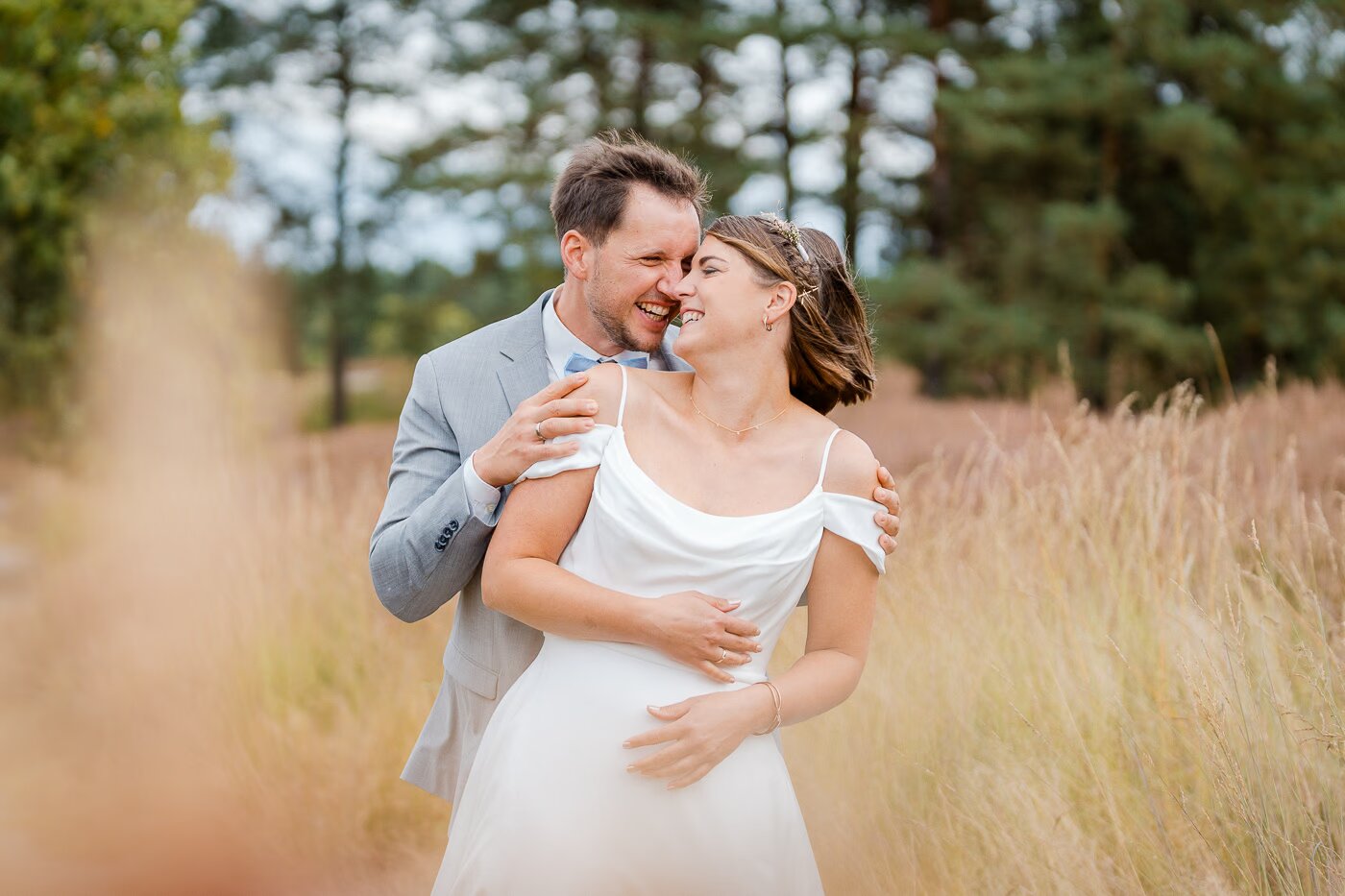 Paarshooting Gifhorner Heide – Brautpaar umarmt sich lachend inmitten hoher Gräser und Kiefern, Bräutigam im hellgrauen Anzug, Braut im fließenden Kleid mit Blumenschmuck im Haar, goldenes Licht, Hochzeitsfotografie Gifhorner Heide Kathrin Skorpel