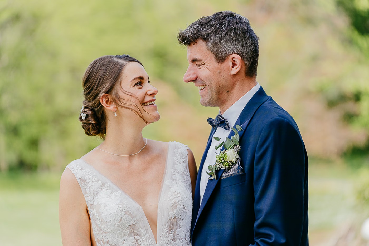 Paarshooting Schlosspark Cremlingen – Brautpaar im Profil lächelt sich liebevoll an, Bräutigam mit gemustertem Fliege und Blumenanstecker, weich verschwommener Grünhintergrund, Hochzeitsfotografie Schlosspark Cremlingen Kathrin Skorpel