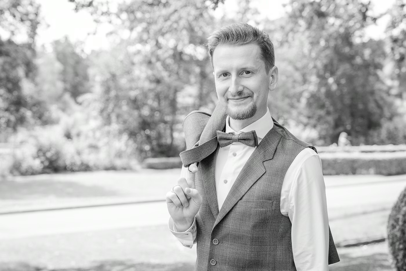 Paarshooting Bad Helmstedt – Schwarz-Weiß-Portrait, Bräutigam hält lächelnd sein Jackett über der Schulter, Weste und Fliege, Parklandschaft im Hintergrund, selbstbewusste Gelassenheit, Hochzeitsfotografie Bad Helmstedt Kathrin Skorpel
