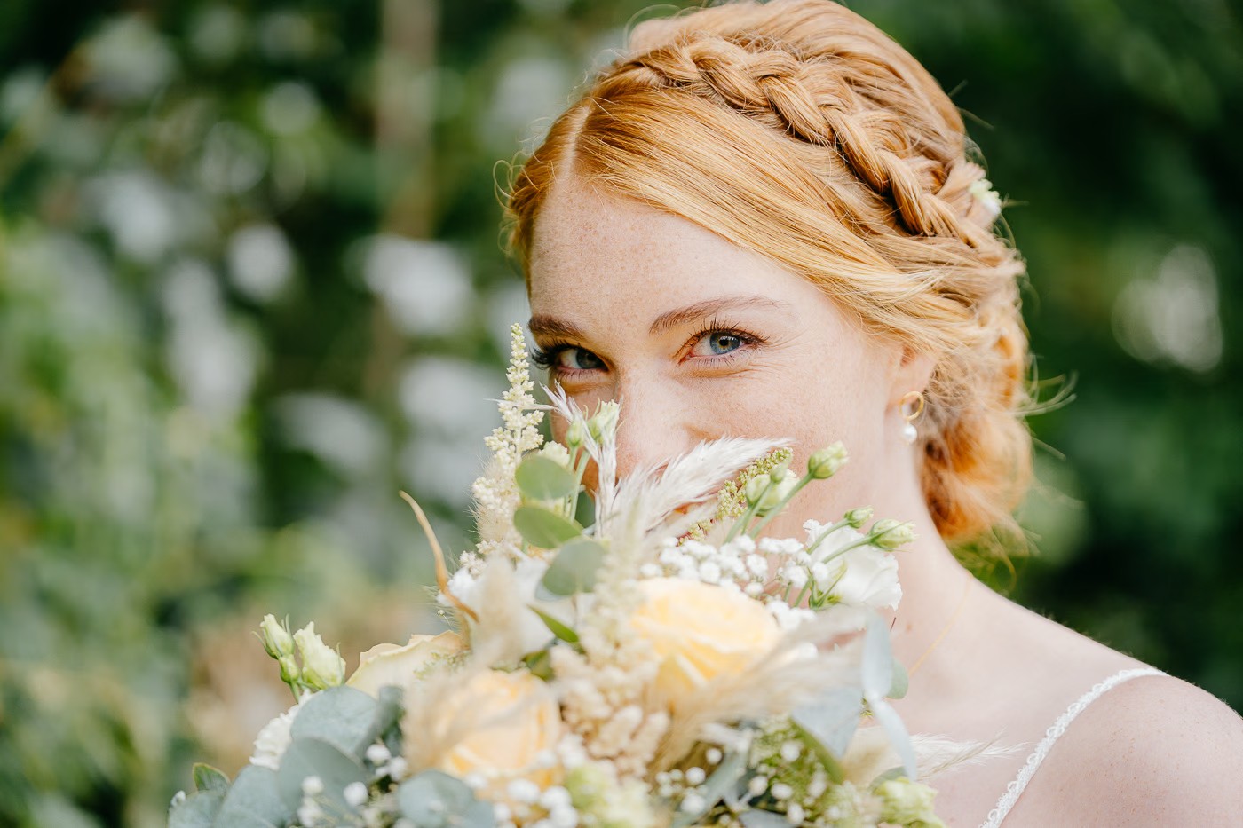 Paarshooting Bad Harzburg – Braut schaut verspielt lächelnd hinter ihrem duftigen Brautstrauß aus Pastellrosen und Trockenblumen hervor, natürliche Ausstrahlung mit Sommersprossen, romantische Waldkulisse, Hochzeitsfotografie Bad Harzburg Kathrin Skorpel