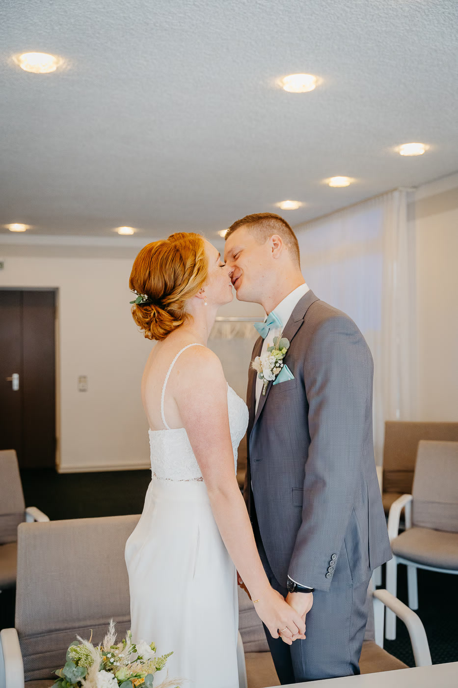 Standesamt Hochzeit Gifhorn – Brautpaar küsst sich stehend im modernen Trauzimmer, Braut mit rotem Dutt und Blüten im Haar, Bräutigam mit hellblauer Fliege, erster Kuss als Eheleute, Hochzeitsreportage Gifhorn Kathrin Skorpel