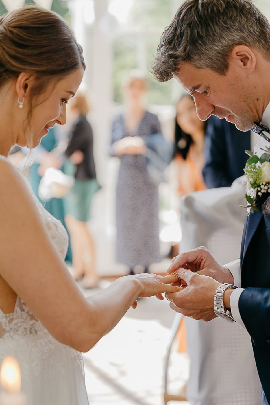 Standesamt Hochzeit Wolfenbüttel – Bräutigam steckt Braut den Ring an den Finger, beide lachen glücklich auf die Hände herab, Gäste stehen im Hintergrund, bewegender Ringtausch, Hochzeitsfotos Wolfenbüttel Kathrin Skorpel