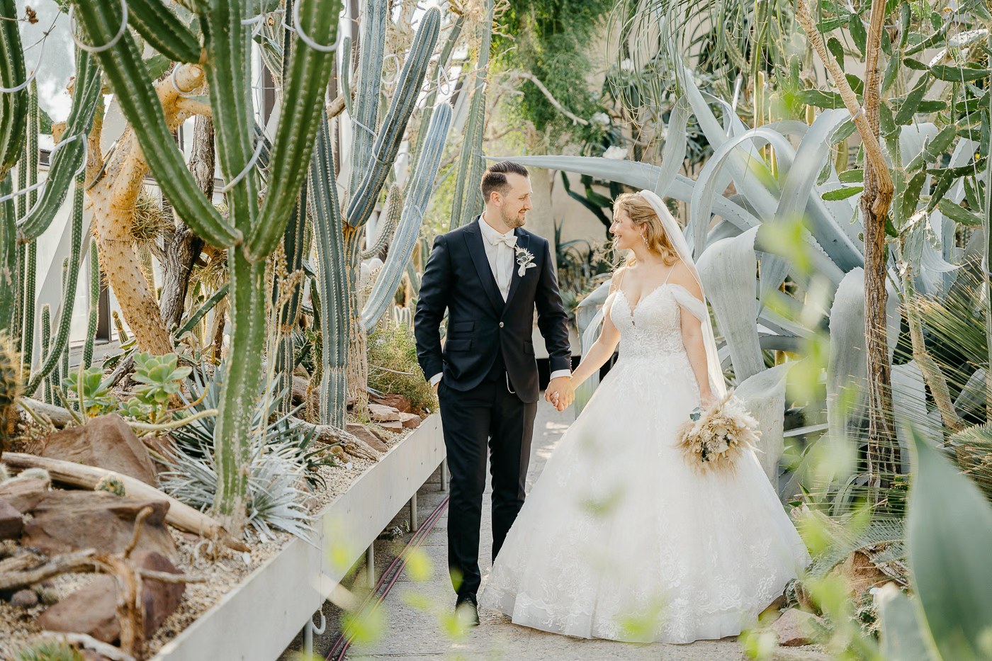 Hochzeitspaarshooting Kakteengarten – Brautpaar Hand in Hand zwischen tropischen Pflanzen, außergewöhnliche Hochzeitsfotos